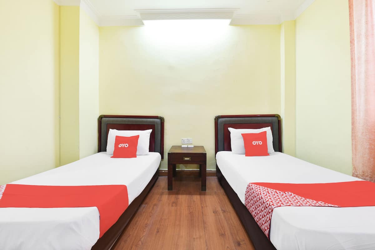 A hotel room with two single beds, each with a red and white bedspread, a small bedside table with a lamp, and a plain wall with a light switch.