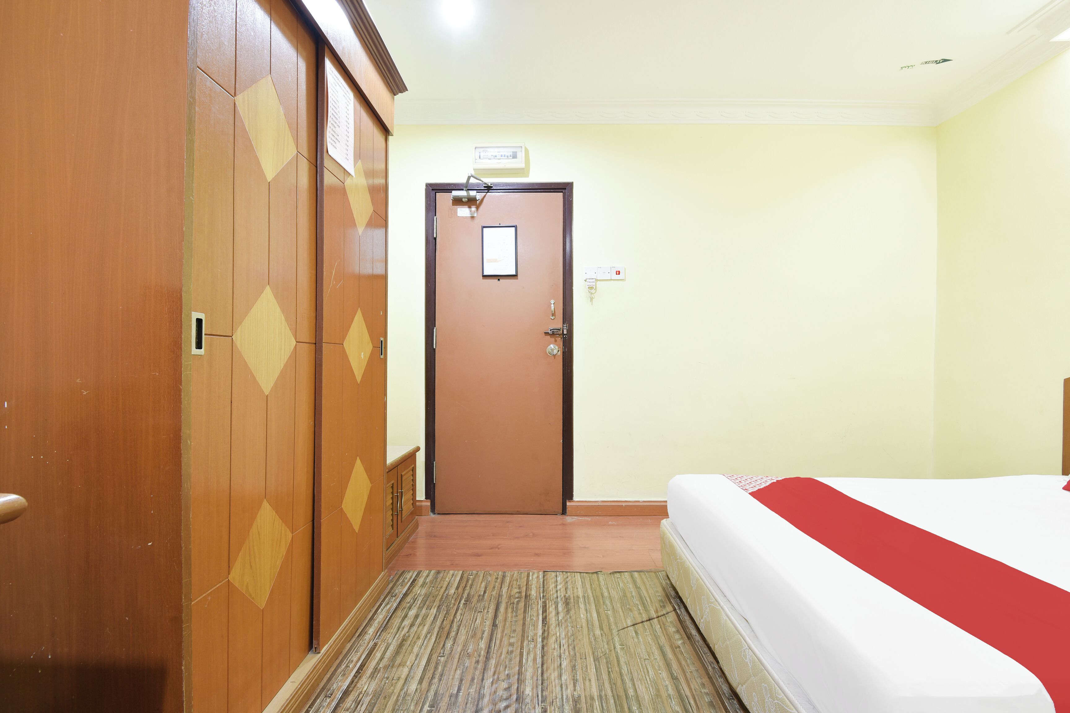 A hotel room with a wooden wardrobe, a door with a sign, a bed with a red blanket, and a carpeted floor.