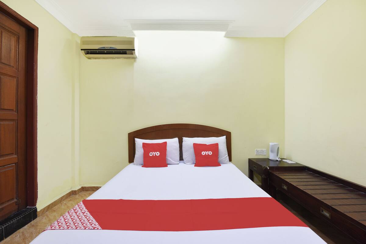 A hotel room with a bed, two red pillows with "OYO" text, a wooden headboard, a bedside table with a phone and a cup, and a window with an air conditioning unit.