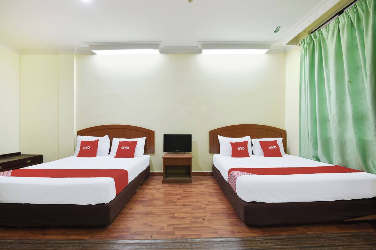A hotel room with two beds, each with white bedding and red pillows, a small bedside table with a TV, and a green curtain on the window.