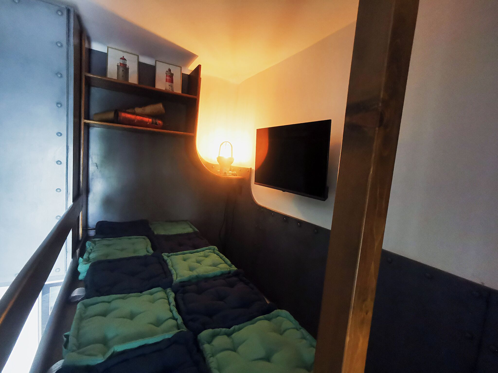 Una cama individual superpuesta con un televisor fijado a la pared, un estante con objetos y una lámpara de pared.