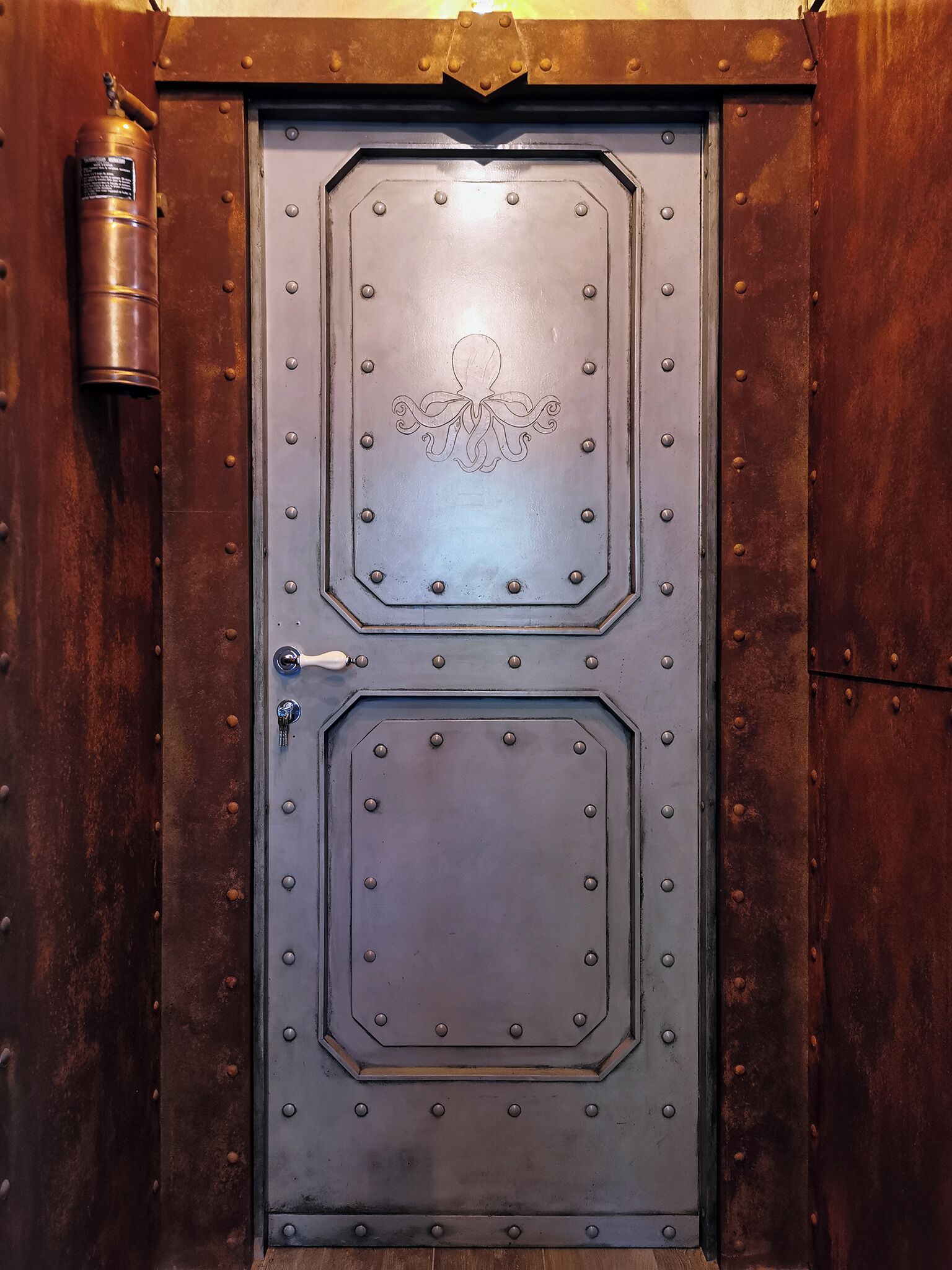 Una puerta metálica grande y octogonal, con manija y cerradura, asegurada con varios cerrojos.