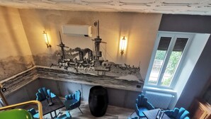 A dining area with blue chairs and a table, a ship mural on the wall, and a window with blinds. - The Kraken Inn (Bergamo)