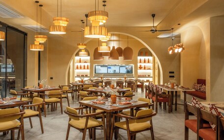 A spacious dining area with multiple tables and chairs, warm lighting, and a large TV screen in the background.