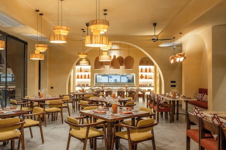 A spacious dining area with multiple tables and chairs, warm lighting, and a large TV screen in the background.