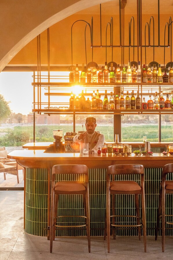 Un espace bar avec un barman, des étagères remplies de bouteilles, des tabourets et une vue sur la verdure à l’extérieur.