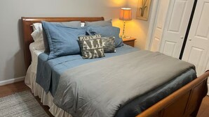 A neatly made bed with blue and grey bedding, a pillow with "PAJAMAS ALL DAY" text, a bedside lamp, and a framed artwork on the wall.
