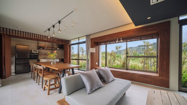 A modern kitchen with a dining area, a sofa with cushions, and large windows offering a view of greenery and hills. - Reflexion Villas GLANZ (Nago)