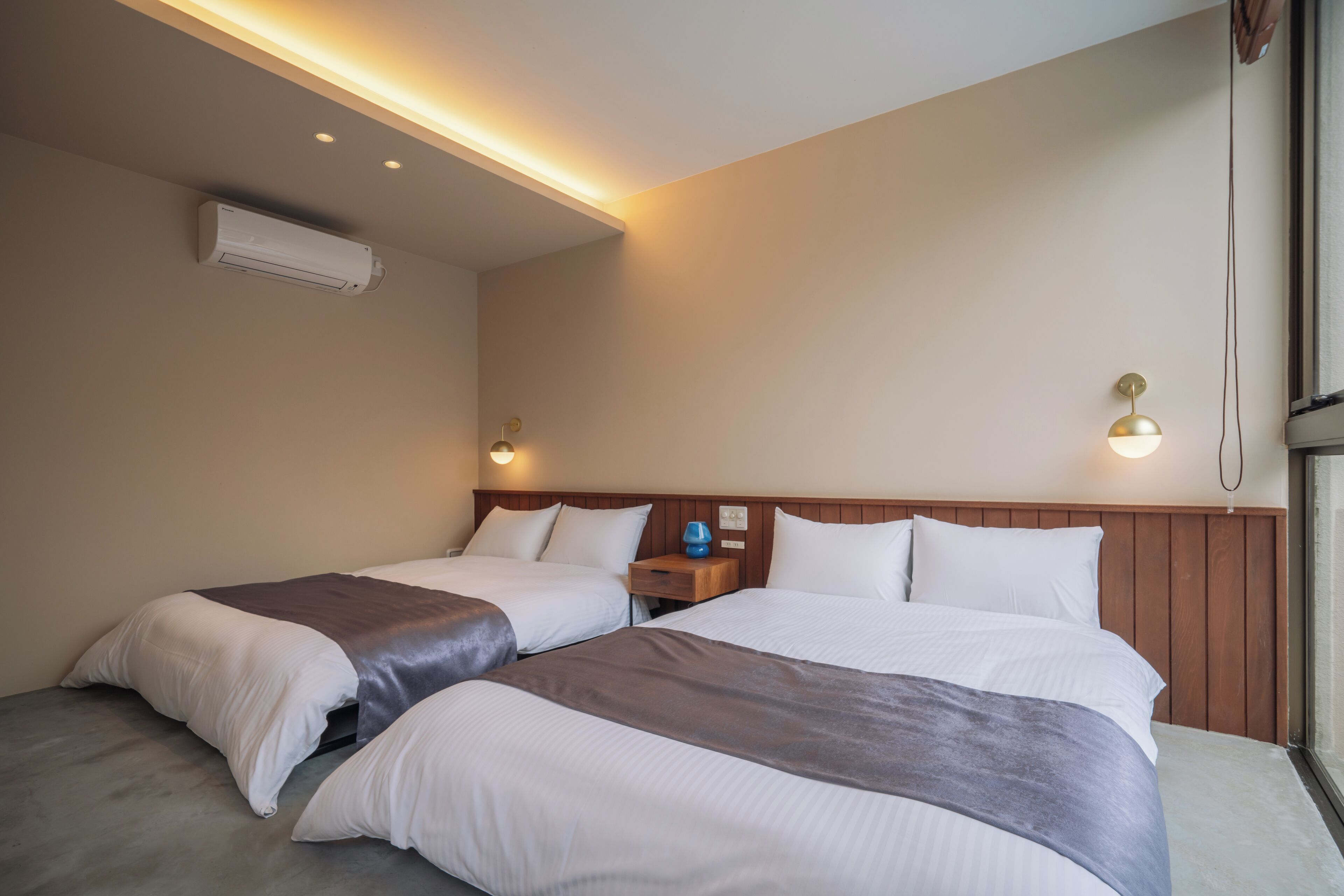 A hotel room with two beds, a wooden headboard, a nightstand with a lamp, and an air conditioning unit.