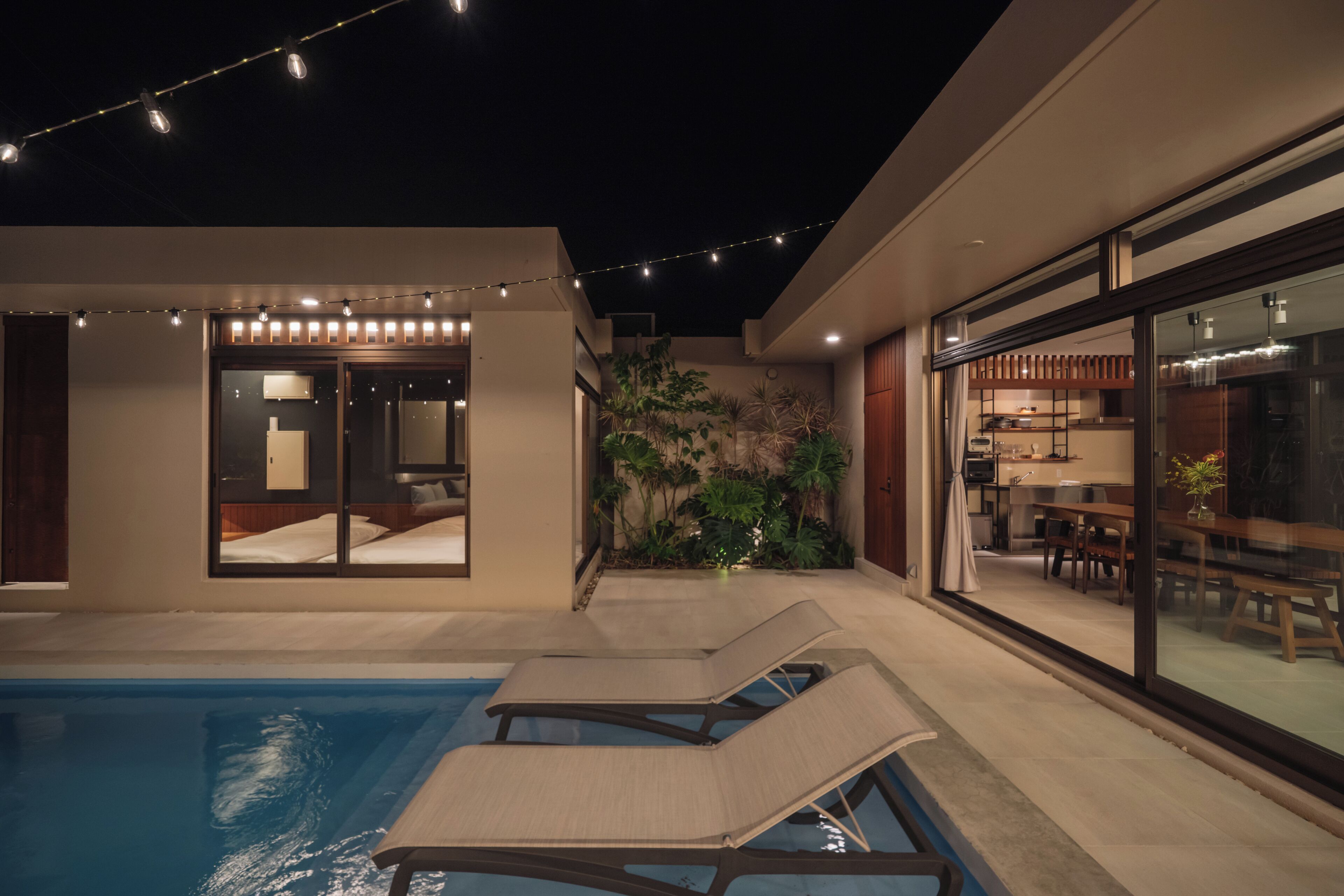 A modern outdoor pool area with lounge chairs, string lights, and a dining area visible through large windows.