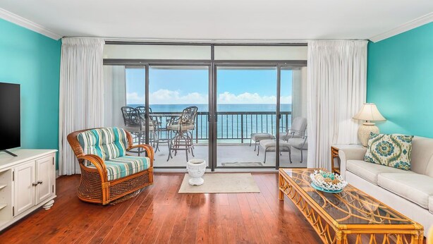 Een kamer met balkon, rieten meubels, een bank, een televisie en uitzicht op zee.