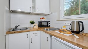 Una cocina con armarios blancos, encimera de madera, un lavamanos, microondas, hervidor y una ventana con vistas a árboles.