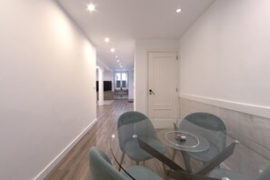 Un couloir moderne et bien éclairé, avec une table et des chaises en verre, qui mène à une porte et offre une vue sur un espace cuisine.