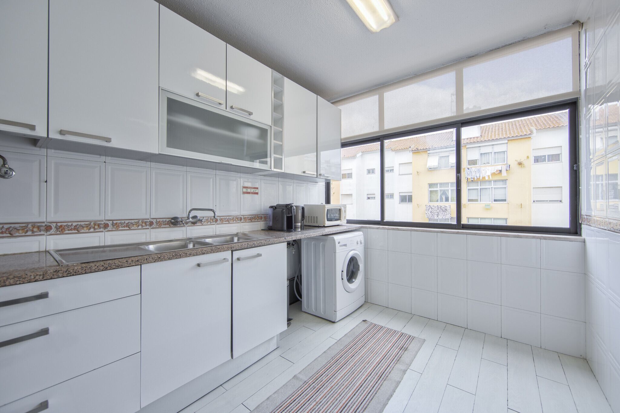 Une cuisine moderne avec des meubles blancs, un lave-linge encastré, un four à micro-ondes et une grande fenêtre donnant sur des immeubles.