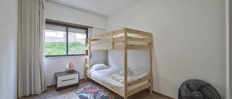 Una habitación con literas, una mesita de noche pequeña con lámpara, un ventilador, una ventana con cortinas y una alfombra estampada.