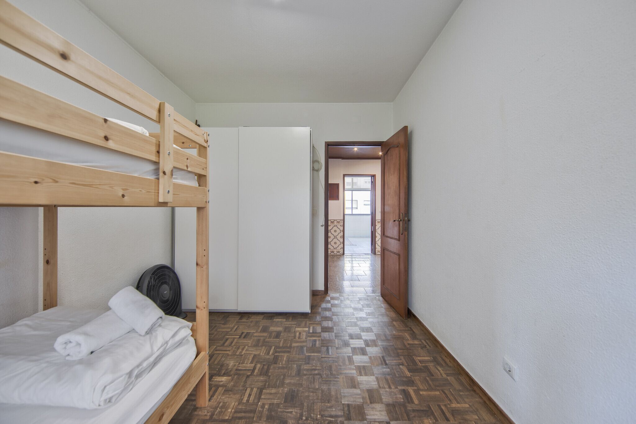 A room with a bunk bed, a white wardrobe, a fan, and a herringbone-patterned floor.