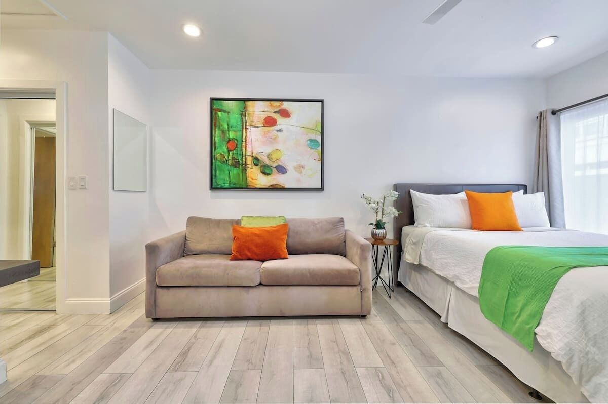 A modern hotel room with a sofa, a bed with a green blanket, and a colorful abstract painting on the wall.