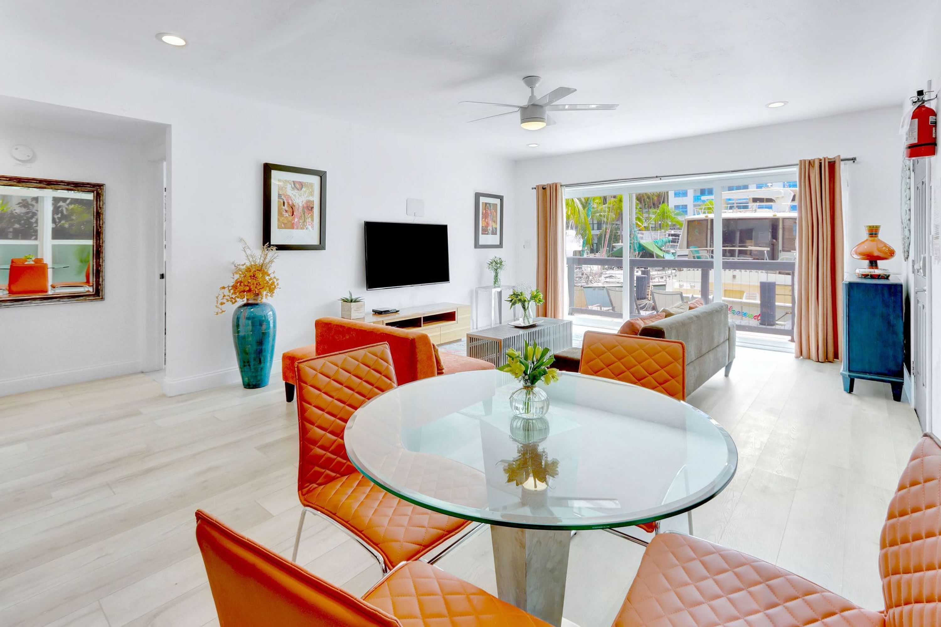 A modern living room with a glass table, orange cushioned chairs, a flat-screen TV, and a large window with a view of the outside.