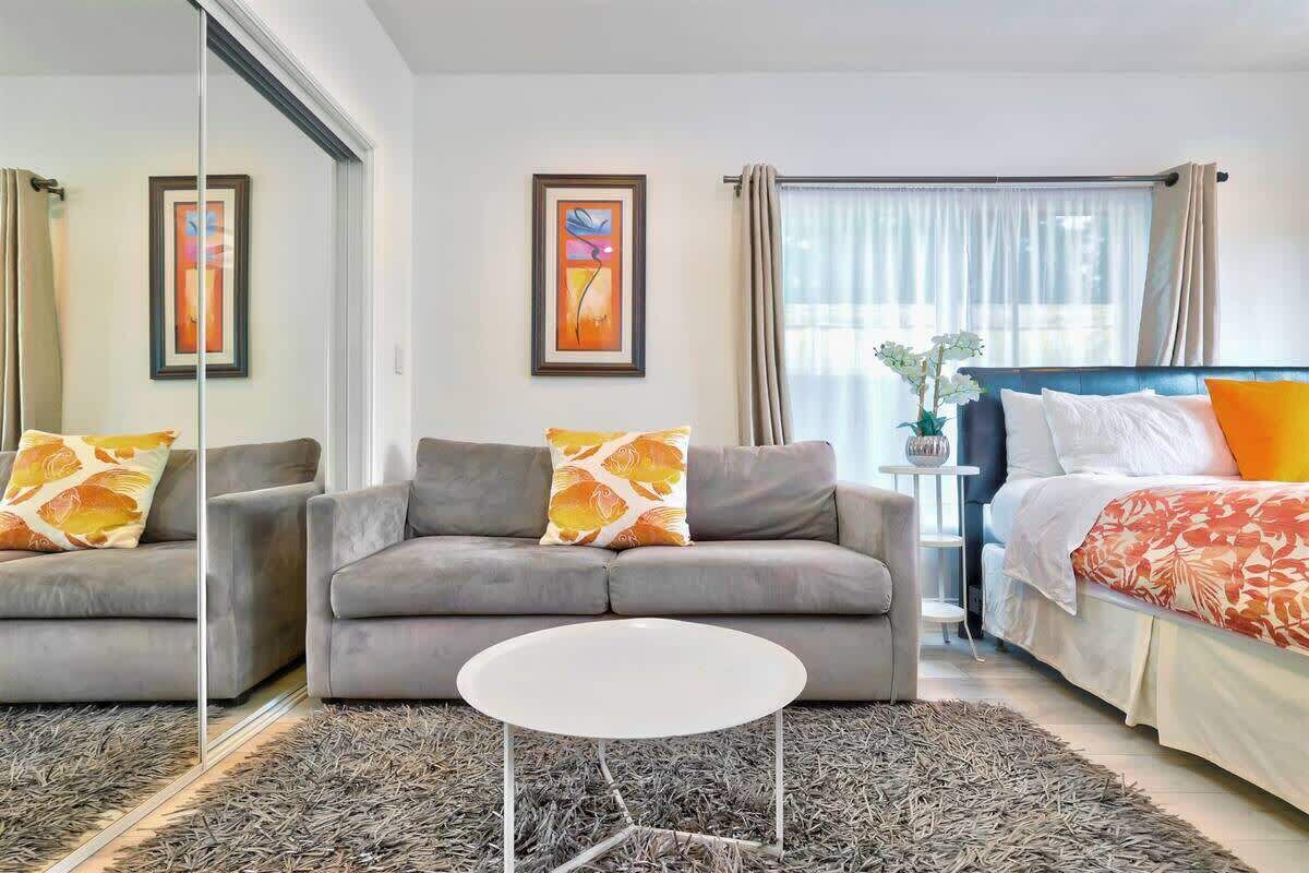 A modern hotel room with a grey sofa, a white coffee table, a bed with a patterned bedspread, and two framed artworks on the walls.
