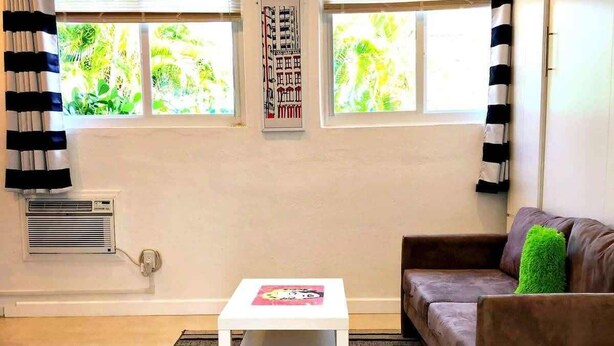 Ein Wohnzimmer mit einem weißen Couchtisch, einem braunen Sofa mit einem grünen Kissen und einem StreifenTeppich. Zwei Fenster mit Vorhängen, eine Klimaanlage und ein an der Wand befestigtes Kunstwerk sind ebenfalls zu sehen.