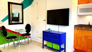 Ein Zimmer mit an der Wand befestigtem Fernseher, einem blauen Schrank mit Schubladen, einem kleinen Tisch mit zwei Stühlen und einem roten Teppich.