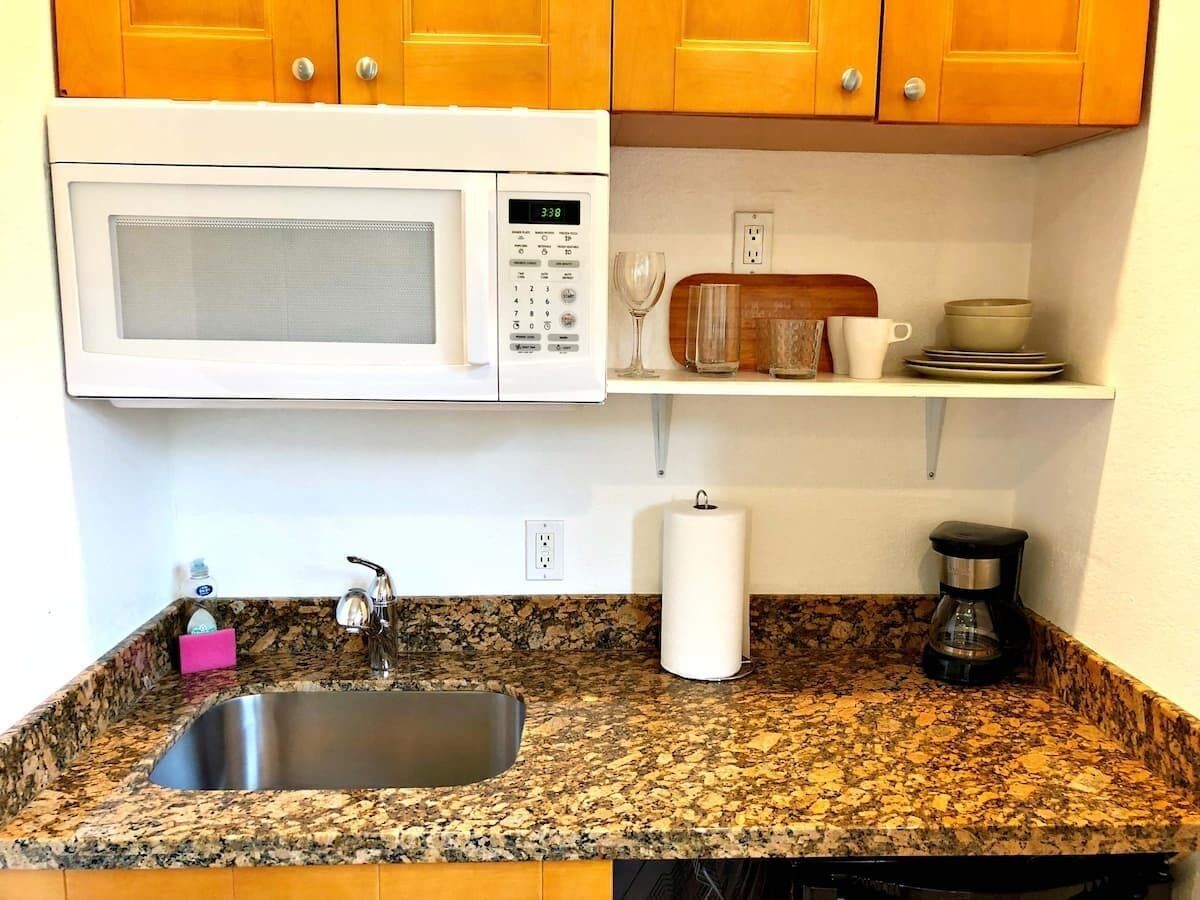 A kitchen with a granite countertop, stainless steel sink, white microwave, and wooden cabinets.