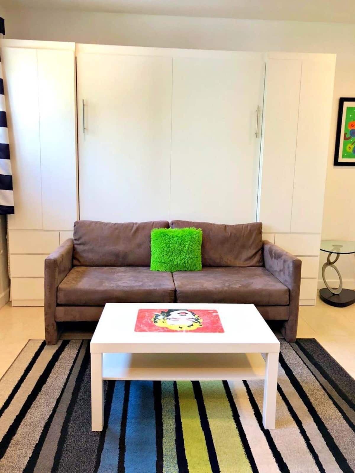 A brown sofa with a green pillow, a white coffee table, and a striped rug in a room with white walls and a framed picture on the wall.
