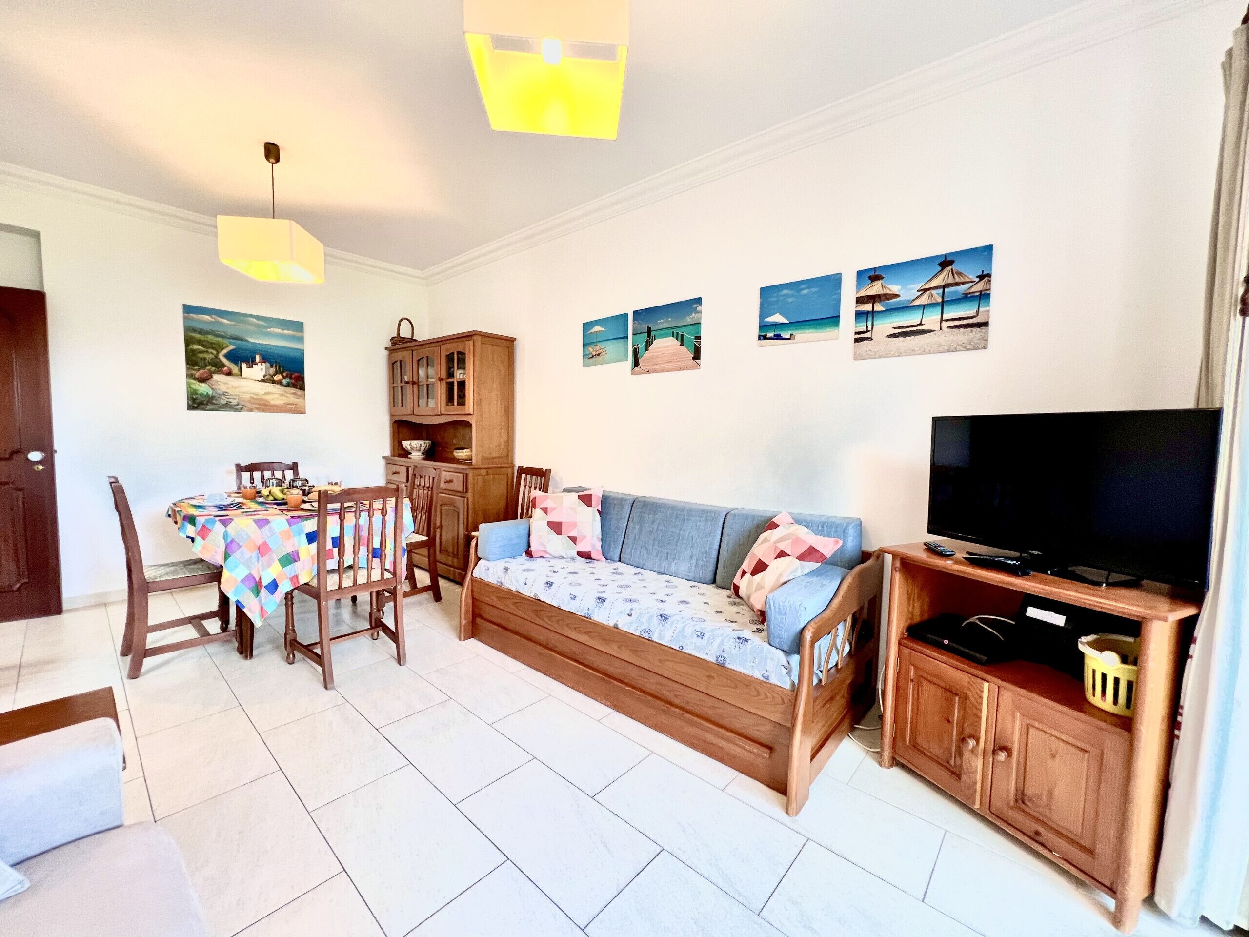 A living room with a sofa, TV, wooden cabinet, dining table, chairs, and wall art.