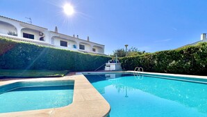 Una piscina azul cristalina de borde curvo, rodeada por un seto cuidado y un edificio con tejado de baldosas y varias ventanas.