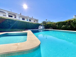 Una piscina azul cristalina de borde curvo, rodeada por un seto cuidado y un edificio con tejado de baldosas y varias ventanas.