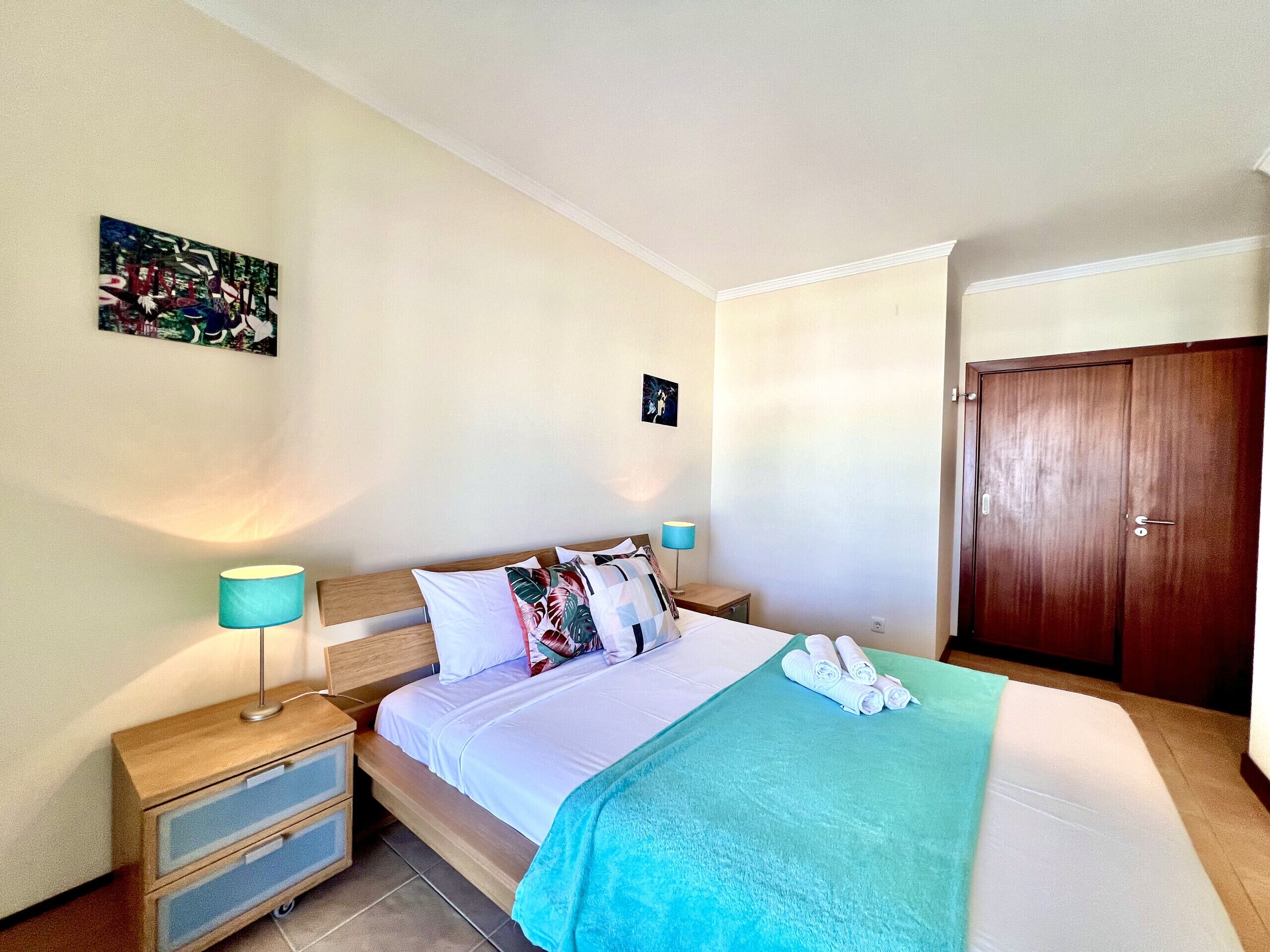 Habitación de hotel con cama, mesitas de noche, lámpara, puerta y un cuadro en la pared.