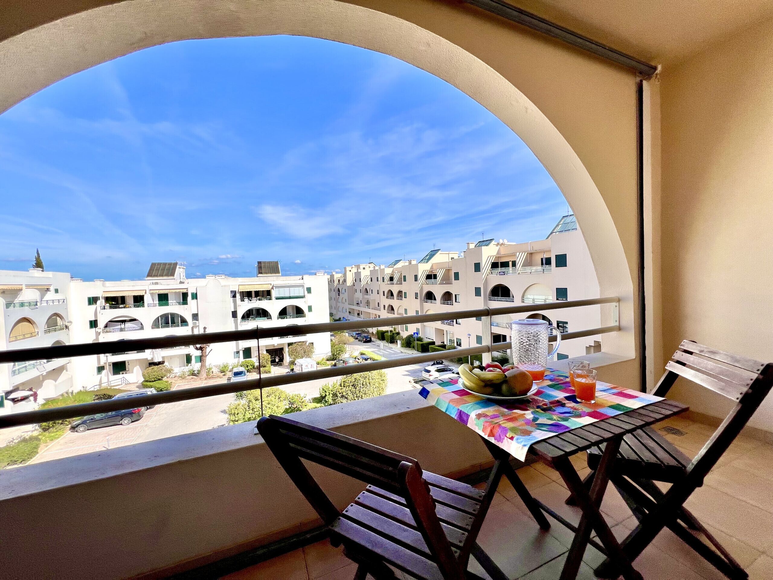 Un balcón con una mesa para dos, con vistas a una zona residencial con edificios blancos y un cielo azul despejado.