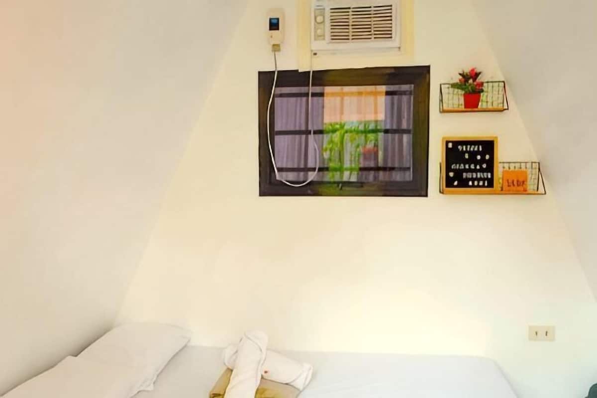 Habitación de hotel con una cama, una ventana con vistas, aire acondicionado de pared y un pequeño estante con una planta y un cartel.