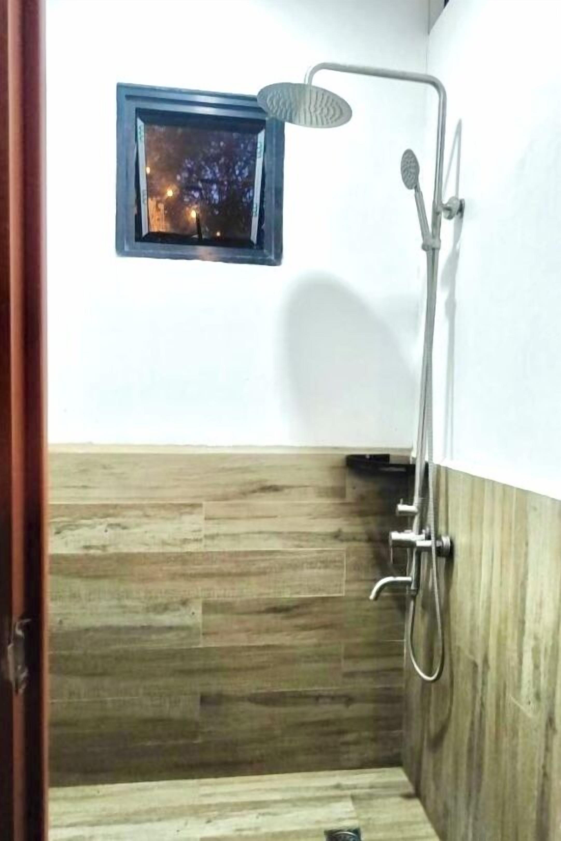 Ein kleines Badezimmer mit Holzboden, einer Dusche mit Duschbrause und einem kleinen Fenster mit Blick auf eine nächtliche Szene.