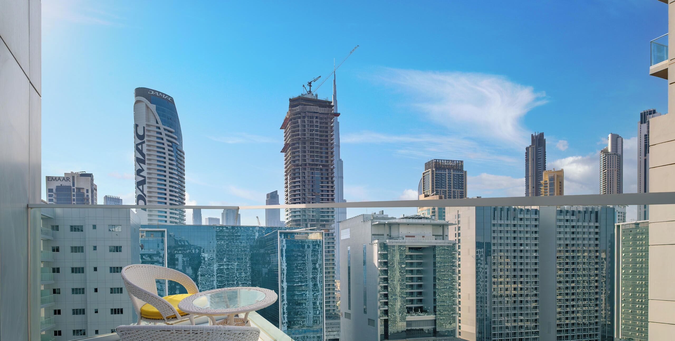 A high-rise building with a balcony featuring a glass table and wicker chairs, overlooking a cityscape with modern architecture.