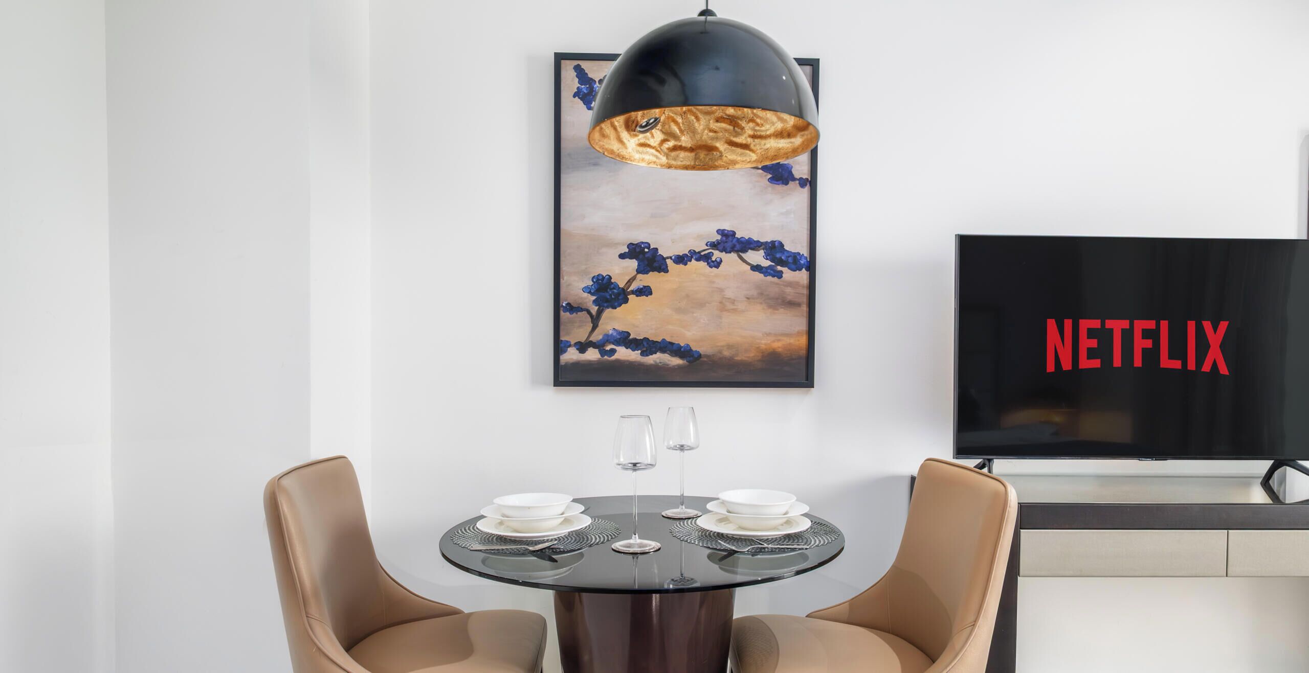 Een moderne eetruimte met een ronde tafel, twee stoelen, een hanglamp en een lijstwerk boven de tafel. Een televisie met het Netflix logo is op de achtergrond te zien.