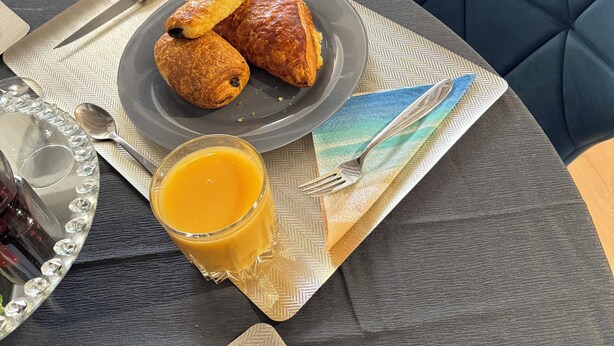 En frukostuppsättning med croissanter, ett glas apelsinjuice och en flaska på ett bord med en mönstrad bordsduk.
