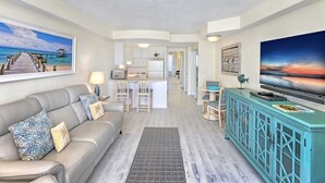 Una sala de estar con sofá, una mesa de centro y televisor. Se observa una cocina al fondo con refrigerador y comedor.