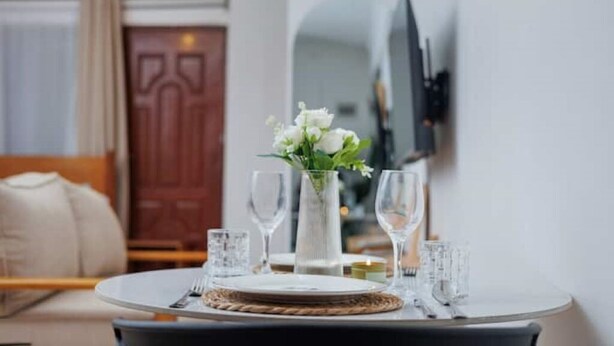 A dining table with a vase of white flowers, wine glasses, and plates, set in a room with a wooden door and a flat-screen TV mounted on the wall.