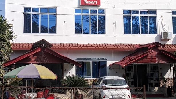 Un bâtiment à deux étages avec une enseigne indiquant « EKORA Resort ». On y trouve des tables et des chaises à l’extérieur, une voiture garée et une zone délimitée par une clôture.