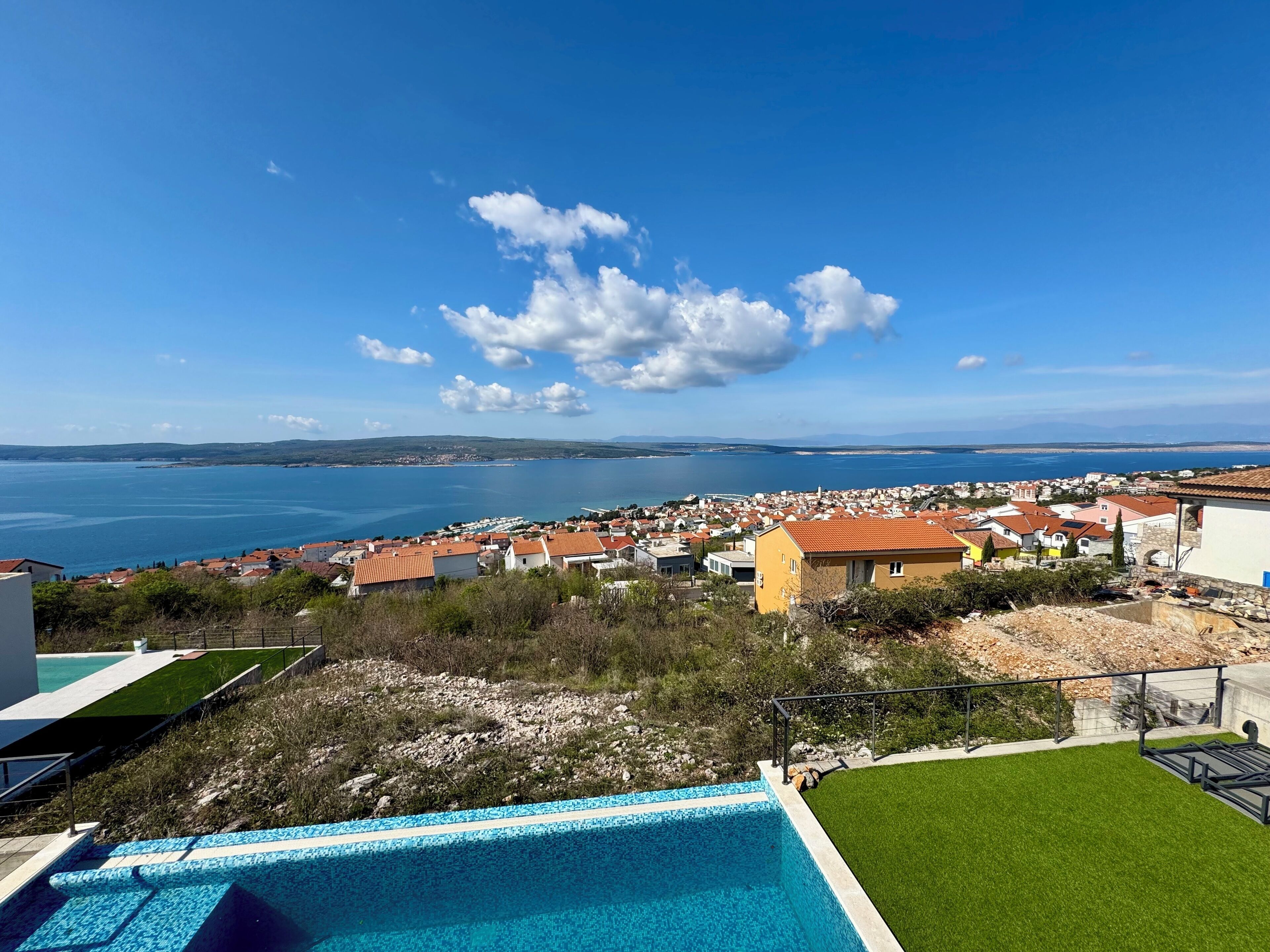 A residential area with a swimming pool, houses with red roofs, and a view of the sea and distant hills.
