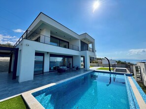 Ett modernt hus i två våningar med en swimmingpool, stora glasfönster och utsikt över havet.