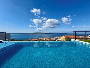 En klar, blå himmel med moln, en swimmingpool med blå kakelplattor och en utsikt över en stad och havet i bakgrunden.