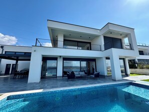 Ett modernt hus i två våningar med en swimmingpool, stora glasfönster och en uteplats.