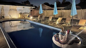Une piscine entourée d’une clôture en bois et d’une maison, avec des chaises longues et des parasols à proximité.
