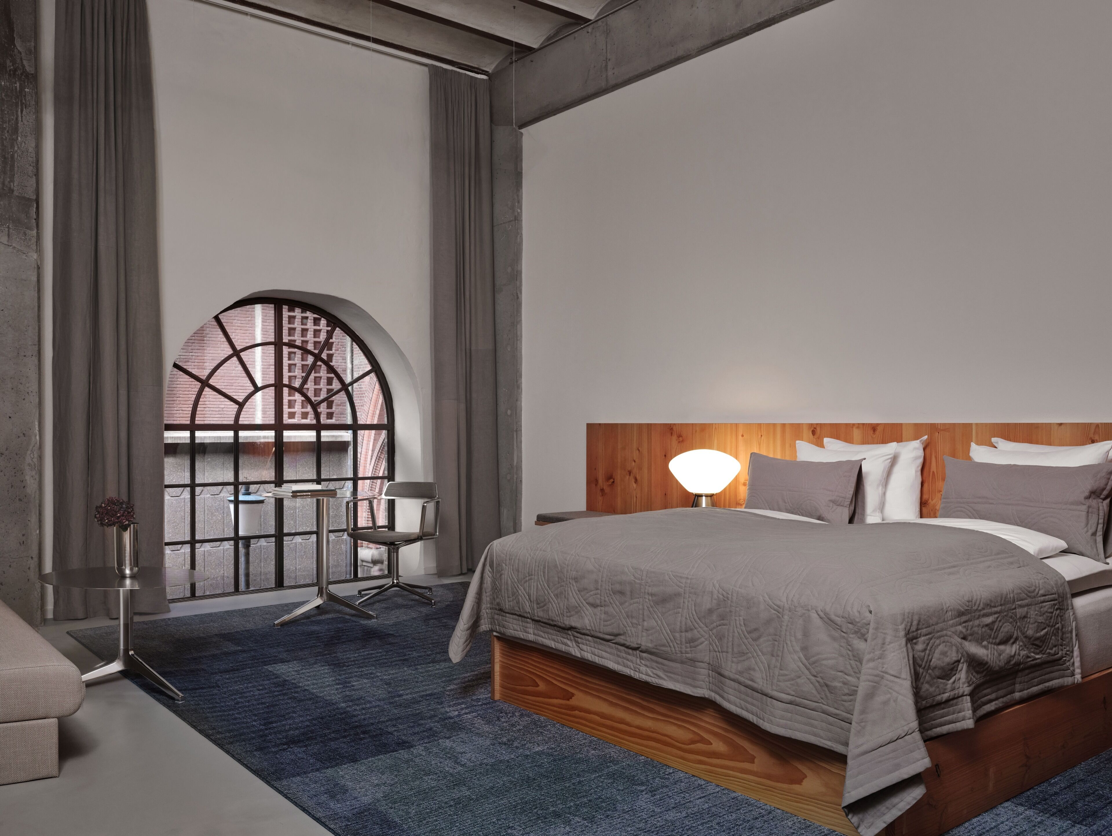 A modern bedroom with a large bed, a wooden headboard, a desk with a chair, and a large arched window.