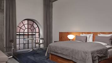 A modern bedroom with a large bed, a wooden headboard, a desk with a chair, and a large arched window.