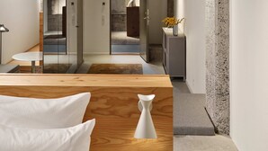 Un interior moderno con techo de concreto, una cama de madera, una puerta de vidrio que da al baño y un mueble pequeño con un florero.