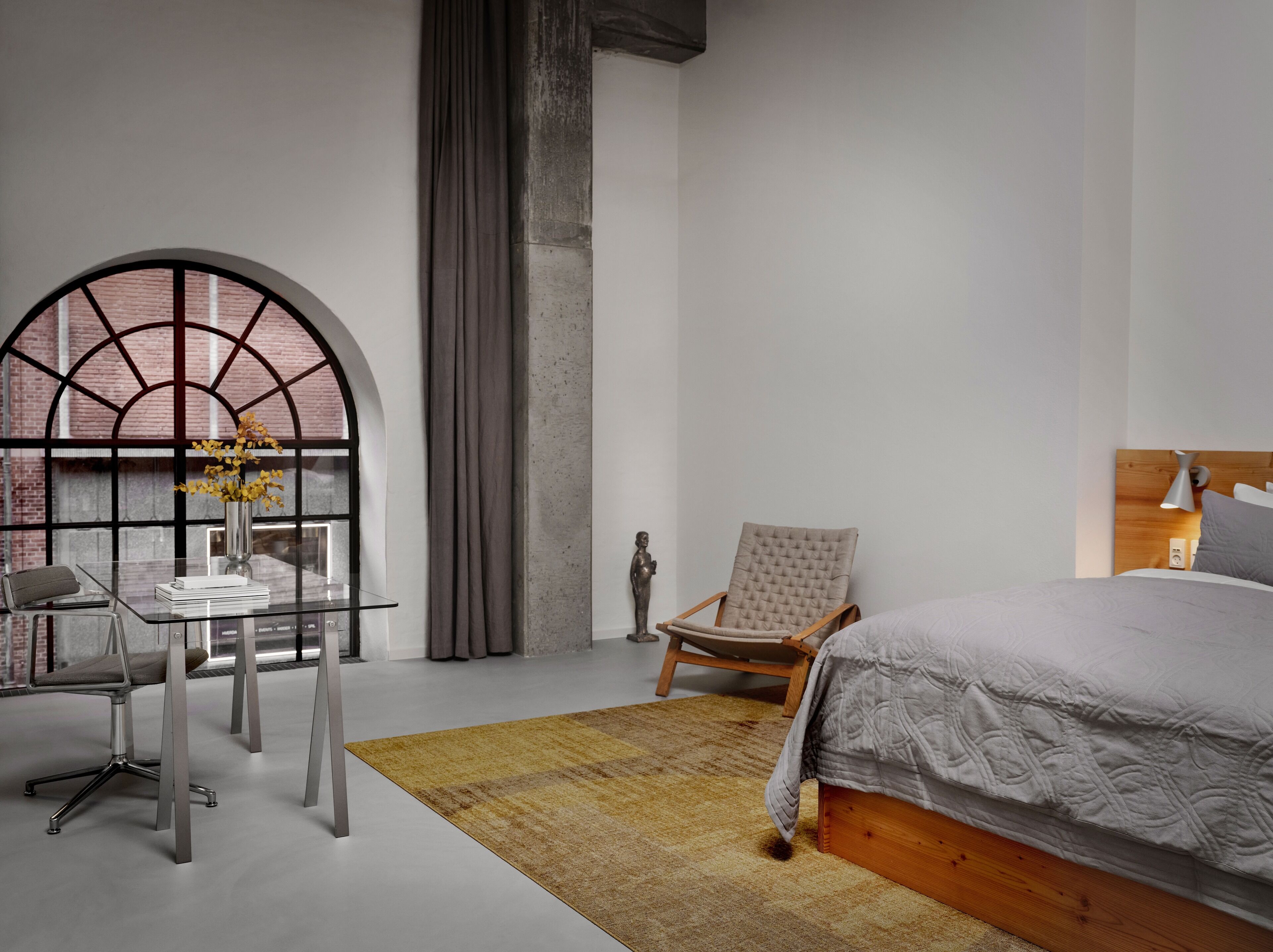 A modern bedroom with a large bed, a desk with a chair, a window with a view of a brick building, and a statue on the floor.
