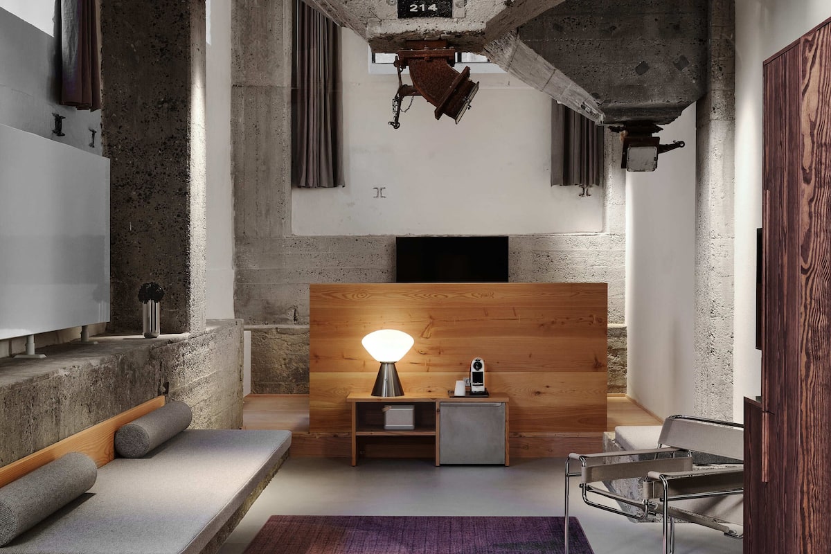 Une chambre moderne avec un plafond en béton, une tête de lit en bois, une petite table de chevet avec une lampe, une chaise en métal et un tapis violet.